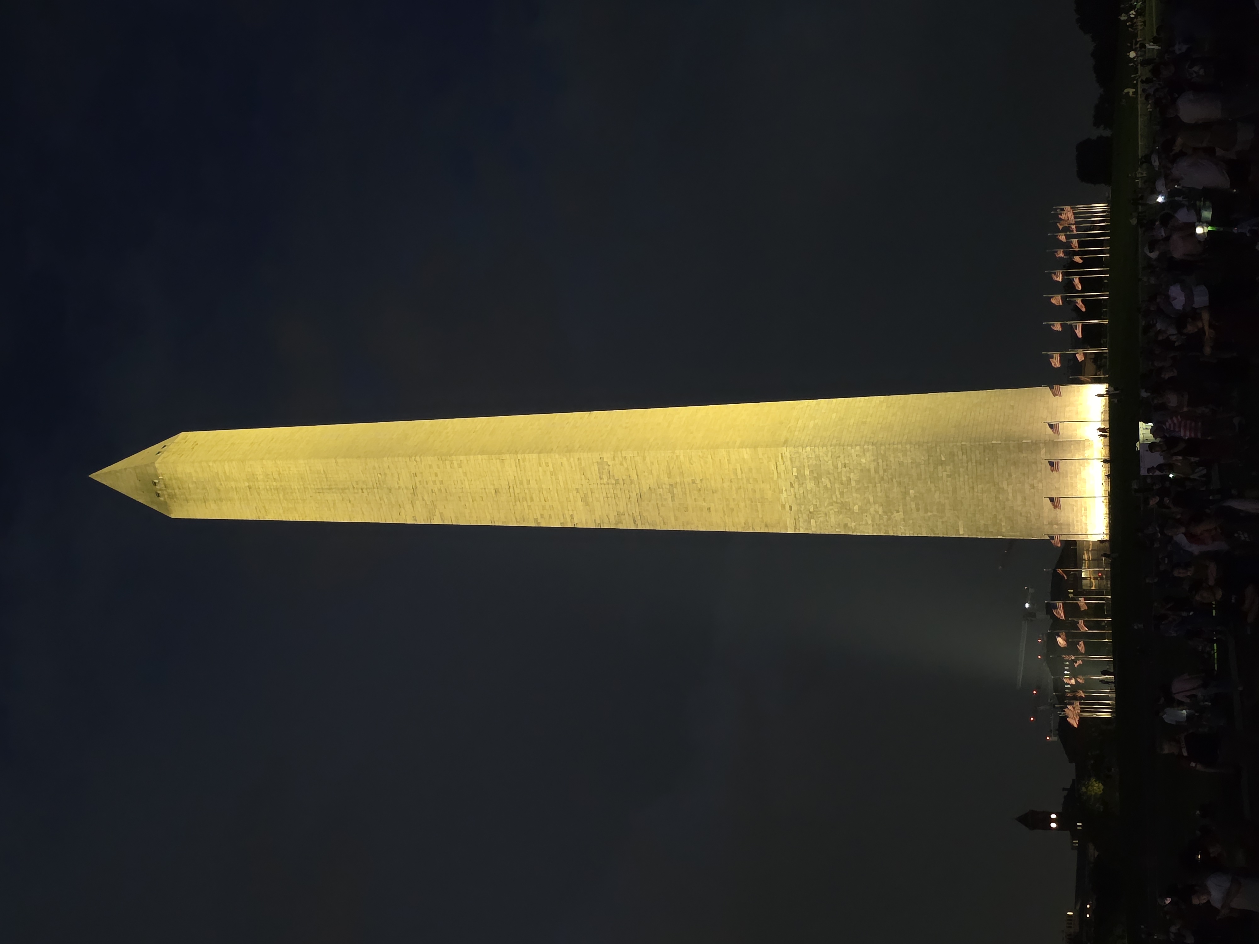 The Washington Monument floodlit at night, ringed by American flags, a crowd gathered at its base.